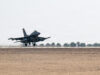Military fighter jet lifting off from a desert runway, afterburners active, with sparse vegetation in the background.
