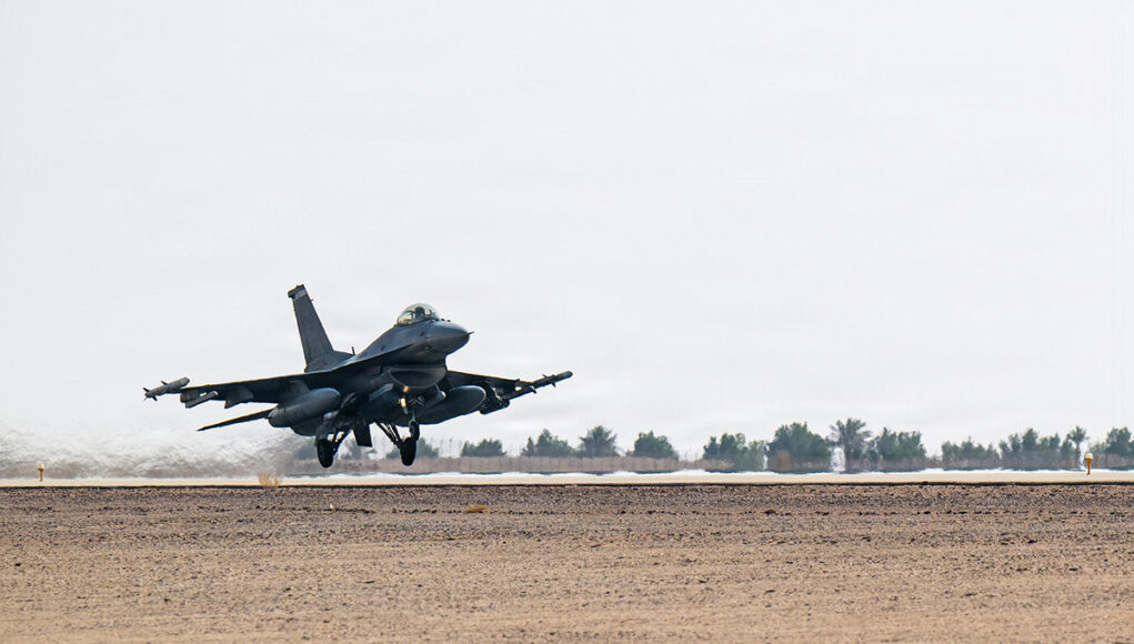Military fighter jet lifting off from a desert runway, afterburners active, with sparse vegetation in the background.