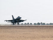 Military fighter jet lifting off from a desert runway, afterburners active, with sparse vegetation in the background.