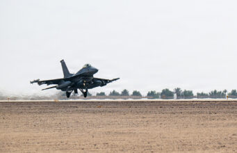 Military fighter jet lifting off from a desert runway, afterburners active, with sparse vegetation in the background.