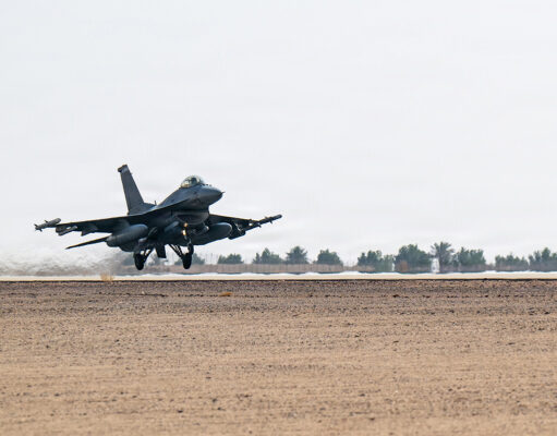 Military fighter jet lifting off from a desert runway, afterburners active, with sparse vegetation in the background.