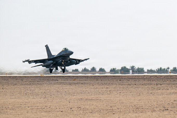 Military fighter jet lifting off from a desert runway, afterburners active, with sparse vegetation in the background.