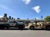 Team LionStrike adds Silverado HD to UK vehicle bid Green pickup truck towing a beige military vehicle along a street with brick buildings on a sunny day after