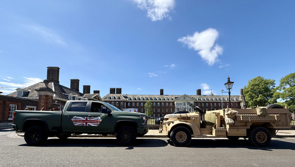 Green pickup truck towing a beige military vehicle along a street with brick buildings on a sunny day after