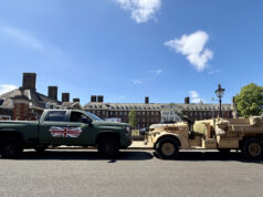 Green pickup truck towing a beige military vehicle along a street with brick buildings on a sunny day after