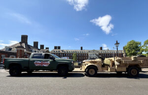 Green pickup truck towing a beige military vehicle along a street with brick buildings on a sunny day after