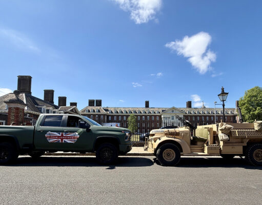 Green pickup truck towing a beige military vehicle along a street with brick buildings on a sunny day after