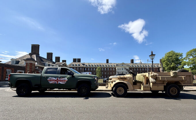 Green pickup truck towing a beige military vehicle along a street with brick buildings on a sunny day after
