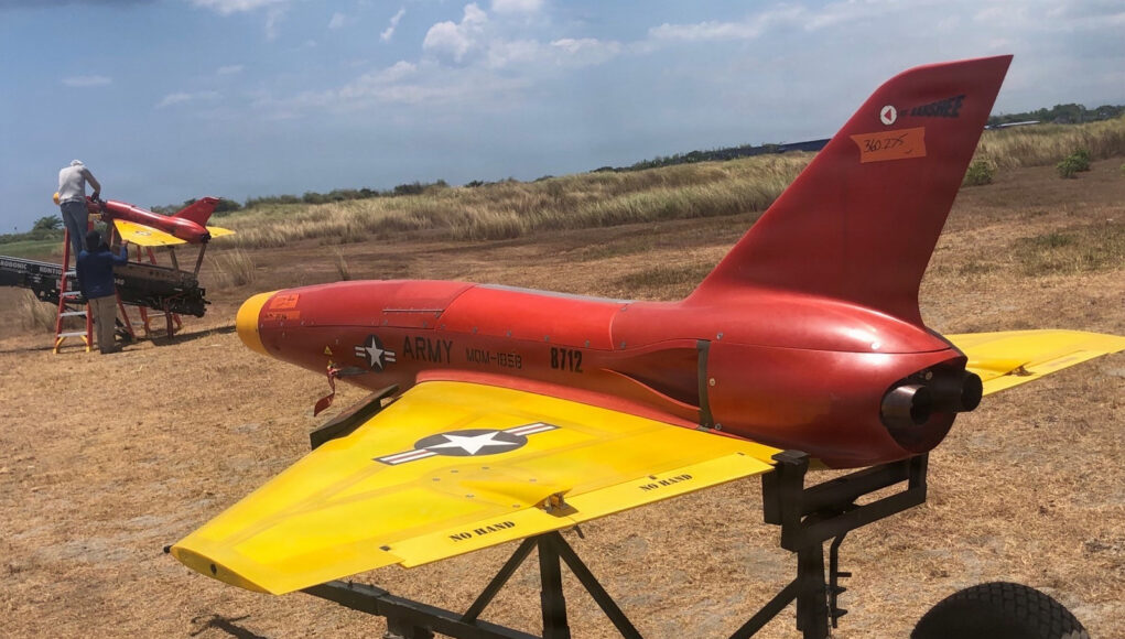 Aerial Drones Elevate IAMD Training for Multinational Forces at Balikatan 2026 Red and yellow military training aircraft mock-up on a wheeled trailer in a dry field, with two technicians working on the nose area far left.