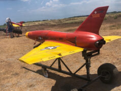 Red and yellow military training aircraft mock-up on a wheeled trailer in a dry field, with two technicians working on the nose area far left.