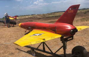 Red and yellow military training aircraft mock-up on a wheeled trailer in a dry field, with two technicians working on the nose area far left.