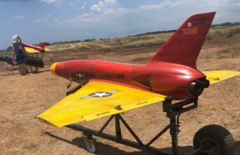 Red and yellow military training aircraft mock-up on a wheeled trailer in a dry field, with two technicians working on the nose area far left.