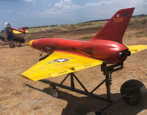 Red and yellow military training aircraft mock-up on a wheeled trailer in a dry field, with two technicians working on the nose area far left.