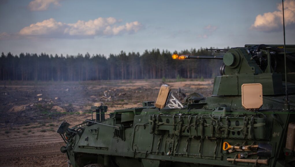 Green armored fighting vehicle firing its main gun in an open field, smoke rising with a forest in the background.