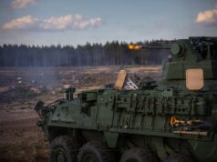 Green armored fighting vehicle firing its main gun in an open field, smoke rising with a forest in the background.