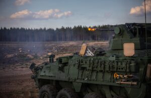 US Stryker opens fire, downs drones in Lithuania Green armored fighting vehicle firing its main gun in an open field, smoke rising with a forest in the background.