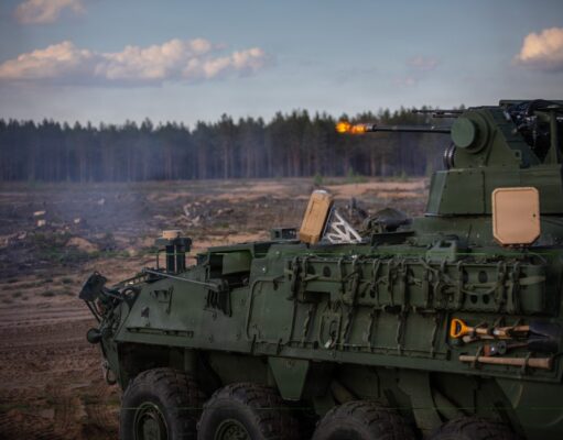 Green armored fighting vehicle firing its main gun in an open field, smoke rising with a forest in the background.