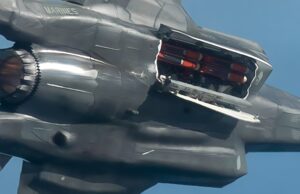 Close-up of a damaged metallic sculpture resembling a fighter jet, with jagged edges and exposed orange interior components against a blue sky.