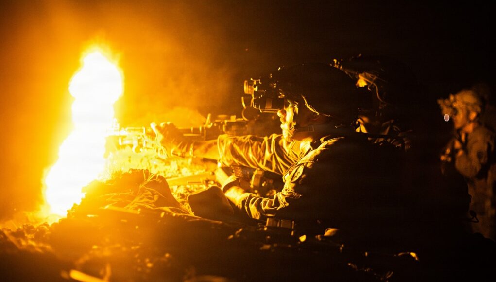 Soldier in night-vision gear firing a rifle beside a large orange flame at night