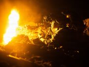 Soldier in night-vision gear firing a rifle beside a large orange flame at night