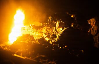 Soldier in night-vision gear firing a rifle beside a large orange flame at night