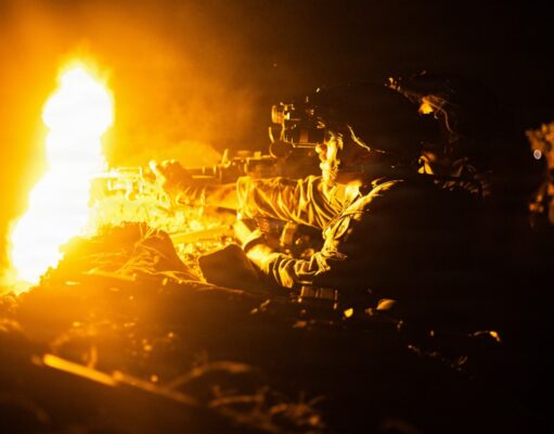 Soldier in night-vision gear firing a rifle beside a large orange flame at night