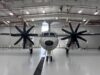 Twin-engine propeller aircraft in a hangar viewed head-on, with red pipes and bright overhead lights above.