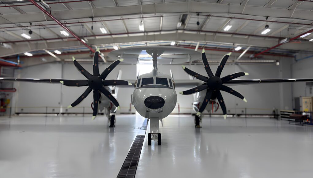 Twin-engine propeller aircraft in a hangar viewed head-on, with red pipes and bright overhead lights above.