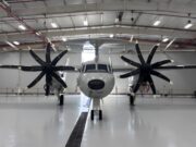 Twin-engine propeller aircraft in a hangar viewed head-on, with red pipes and bright overhead lights above.