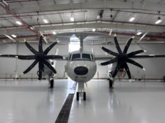 Twin-engine propeller aircraft in a hangar viewed head-on, with red pipes and bright overhead lights above.
