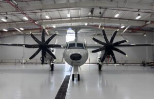 Twin-engine propeller aircraft in a hangar viewed head-on, with red pipes and bright overhead lights above.