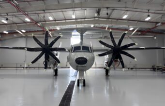 Twin-engine propeller aircraft in a hangar viewed head-on, with red pipes and bright overhead lights above.
