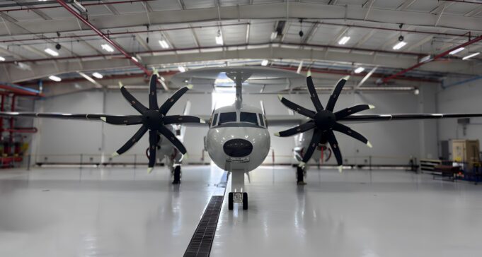 Twin-engine propeller aircraft in a hangar viewed head-on, with red pipes and bright overhead lights above.