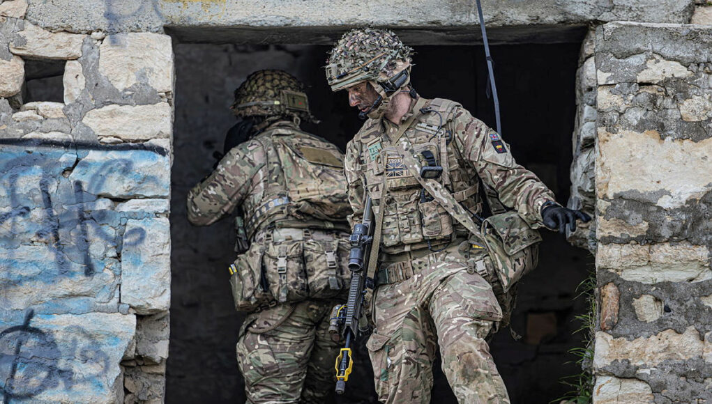 Two soldiers in full combat gear exit a ruined stone building, rifles at the ready.