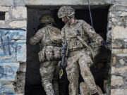 Two soldiers in full combat gear exit a ruined stone building, rifles at the ready.