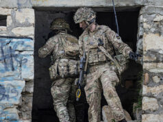 Two soldiers in full combat gear exit a ruined stone building, rifles at the ready.