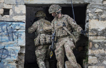 Two soldiers in full combat gear exit a ruined stone building, rifles at the ready.