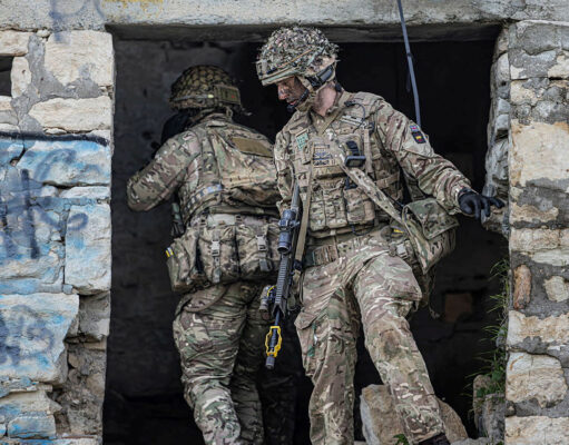 Two soldiers in full combat gear exit a ruined stone building, rifles at the ready.
