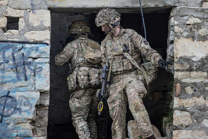 Two soldiers in full combat gear exit a ruined stone building, rifles at the ready.