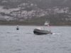 Metal patrol boat with the number 16 speeds across the water, with a rocky hillside in the distance.