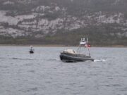 Metal patrol boat with the number 16 speeds across the water, with a rocky hillside in the distance.