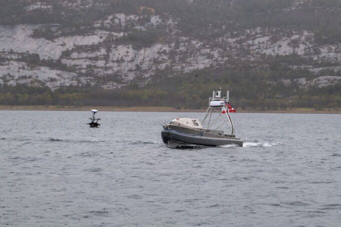Metal patrol boat with the number 16 speeds across the water, with a rocky hillside in the distance.