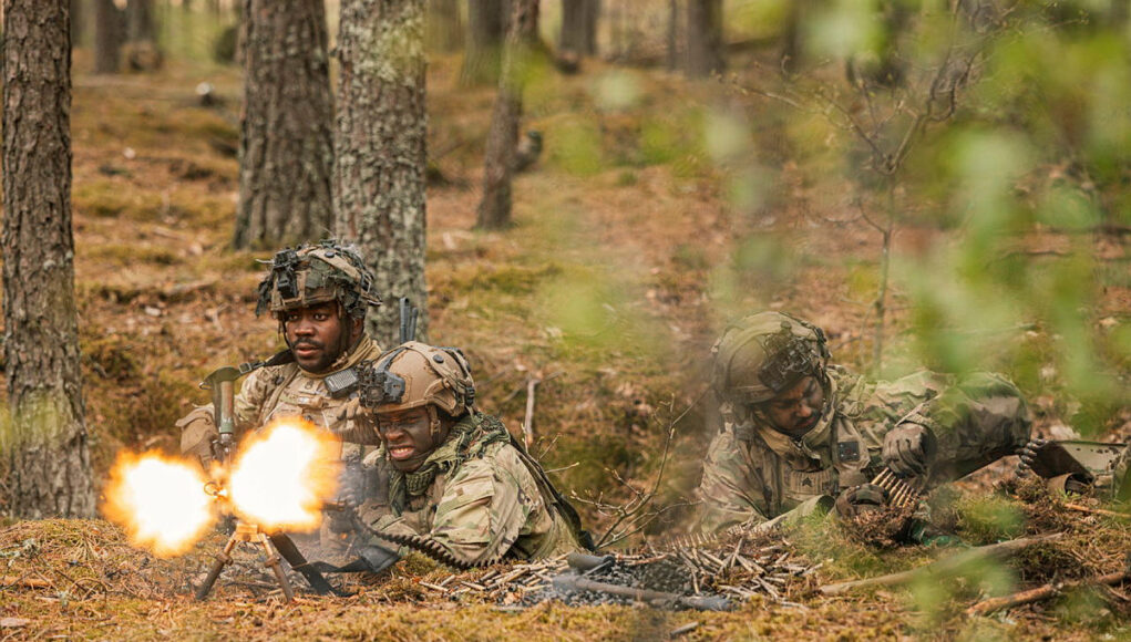 Soldiers in camouflage firing a light machine gun in a forest, muzzle flash brightening the scene.