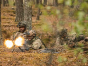 Soldiers in camouflage firing a light machine gun in a forest, muzzle flash brightening the scene.