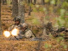 Soldiers in camouflage firing a light machine gun in a forest, muzzle flash brightening the scene.