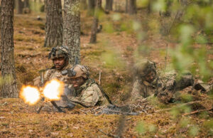 Soldiers in camouflage firing a light machine gun in a forest, muzzle flash brightening the scene.