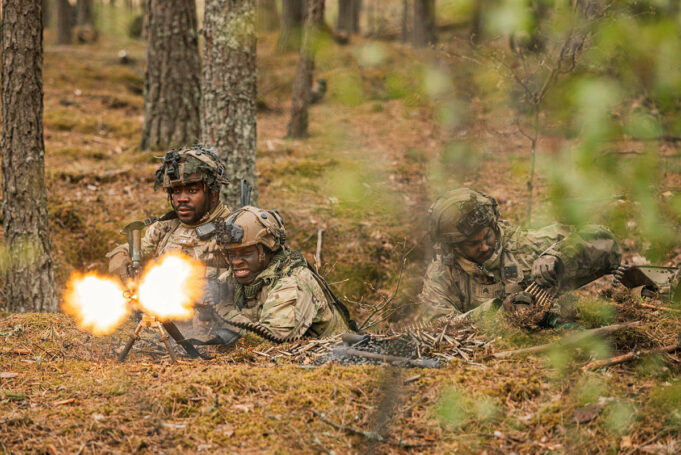 Soldiers in camouflage firing a light machine gun in a forest, muzzle flash brightening the scene.