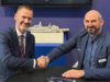 Two men in blue suits shake hands across a table, smiling, with a model ship on a shelf behind them.