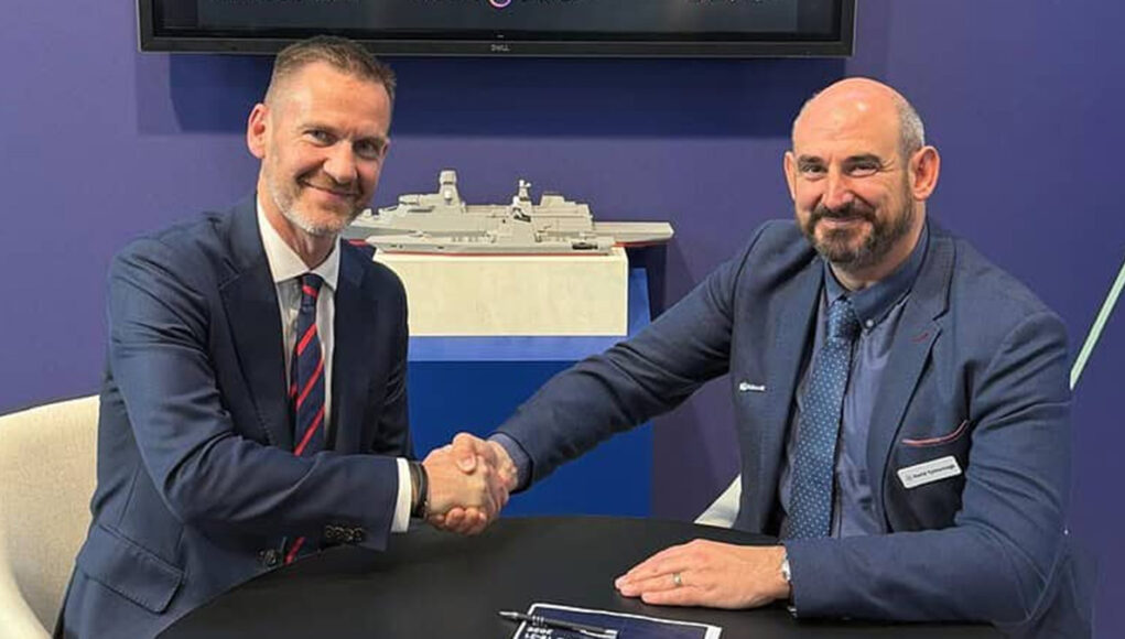 Two men in blue suits shake hands across a table, smiling, with a model ship on a shelf behind them.