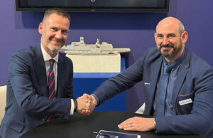 Babcock signs five-year engineering deal with AtkinsRealis Two men in blue suits shake hands across a table, smiling, with a model ship on a shelf behind them.